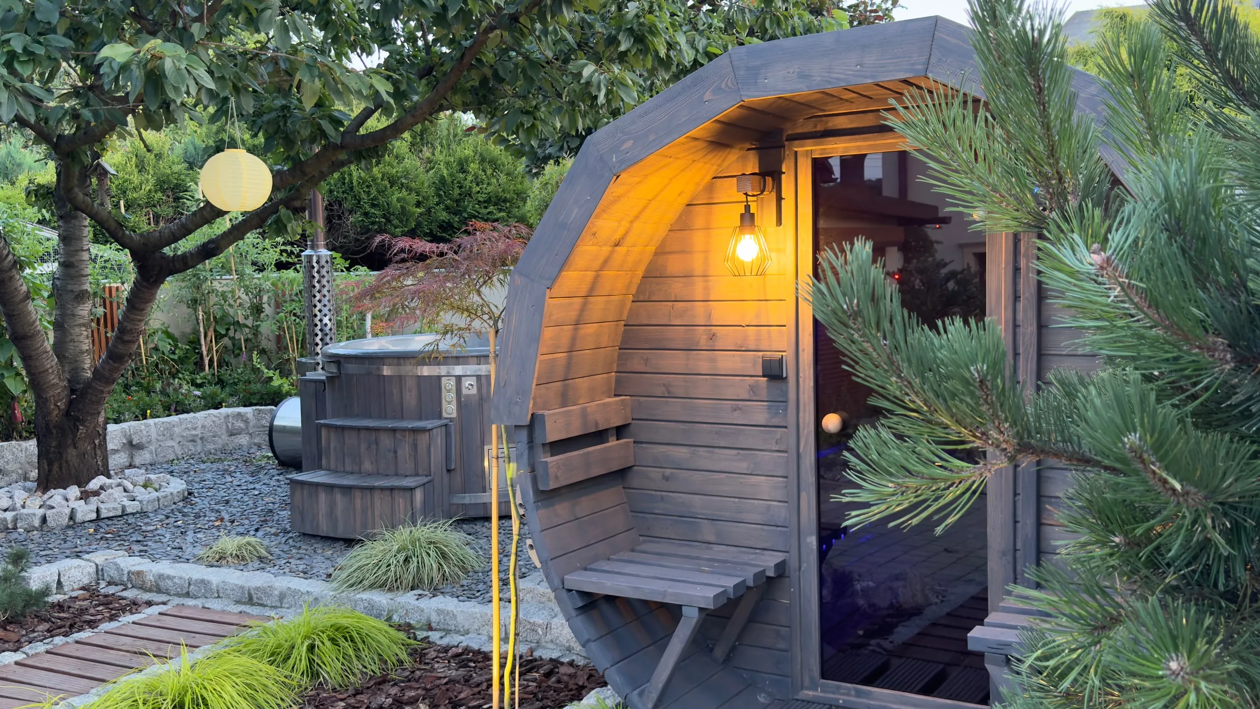 Sauna ogrodowa beczka z zapaloną lampą przy wejściu, w tle balia ogrodowa. Showroom Rozgrzani w Suszcu